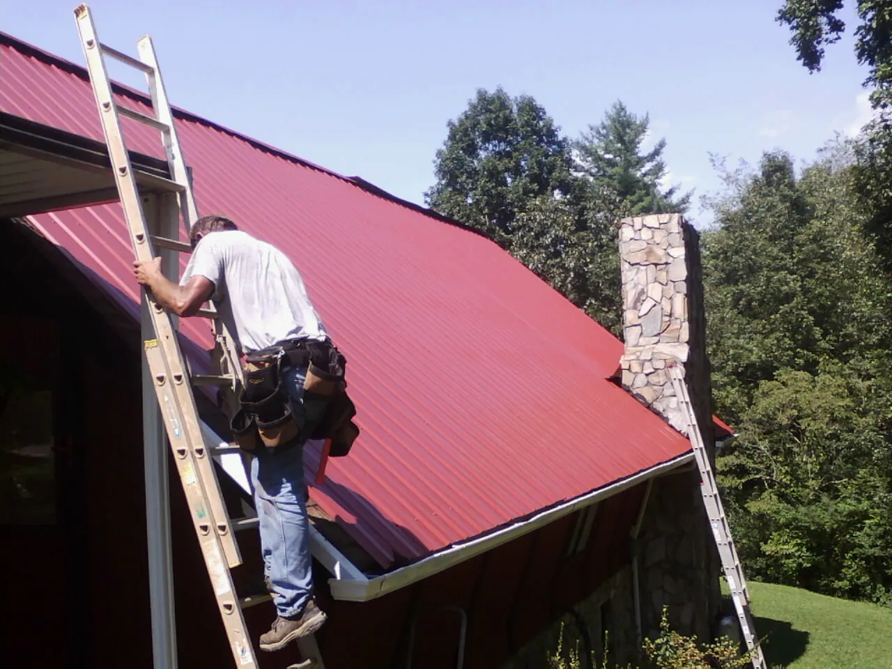 Roofer on a ladder beside a red metal roof and stone chimney — Soffit & Fascia Repair by Roofing Brothers in Whately